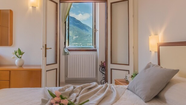 A bedroom with a bed, a nightstand, a dresser, a window with a mountain view, and a vase with flowers. - Casa Greppi Varenna in Varenna (Varenna)
