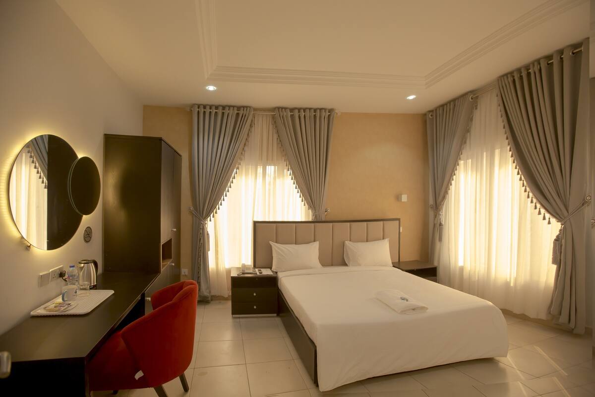 A hotel room with a bed, a desk with a chair, a mirror, a nightstand, and curtains.