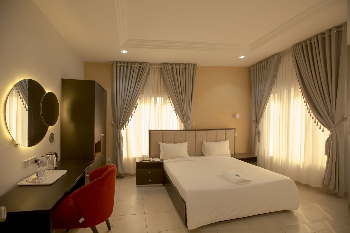 A hotel room with a bed, a desk with a chair, a mirror, a nightstand, and curtains.