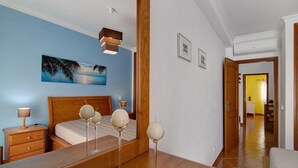 A hotel room with a bed, bedside tables, a mirror, a wall with a painting, and a hallway leading to another room. - Apartment 'Algarvio Ao Sol' with Balcony, Wi-Fi and Air Conditioning (Conceição de Tavira)