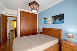 A bedroom with a bed, wooden wardrobe, nightstand, and a wall-mounted lamp.