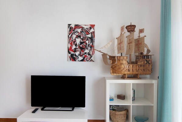 Un televisor de pantalla plana sobre un soporte blanco, un modelo de barco de madera y un cuadro abstracto geométrico en la pared.
