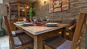 Un comedor con una mesa de madera dispuesta para cuatro personas, rodeada de sillas acolchadas. Un jarrón grande con flores está sobre la mesa, y hay una pared de piedra con un cuadro colgado.