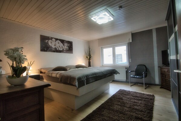 Un dormitorio con cama, escritorio, silla, ventana y un cuadro en la pared.