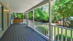Eine überdachte Veranda mit Holzdeck, weißen Geländern und Blick auf einen grünen Garten mit Bäumen und einem Holzzaun.