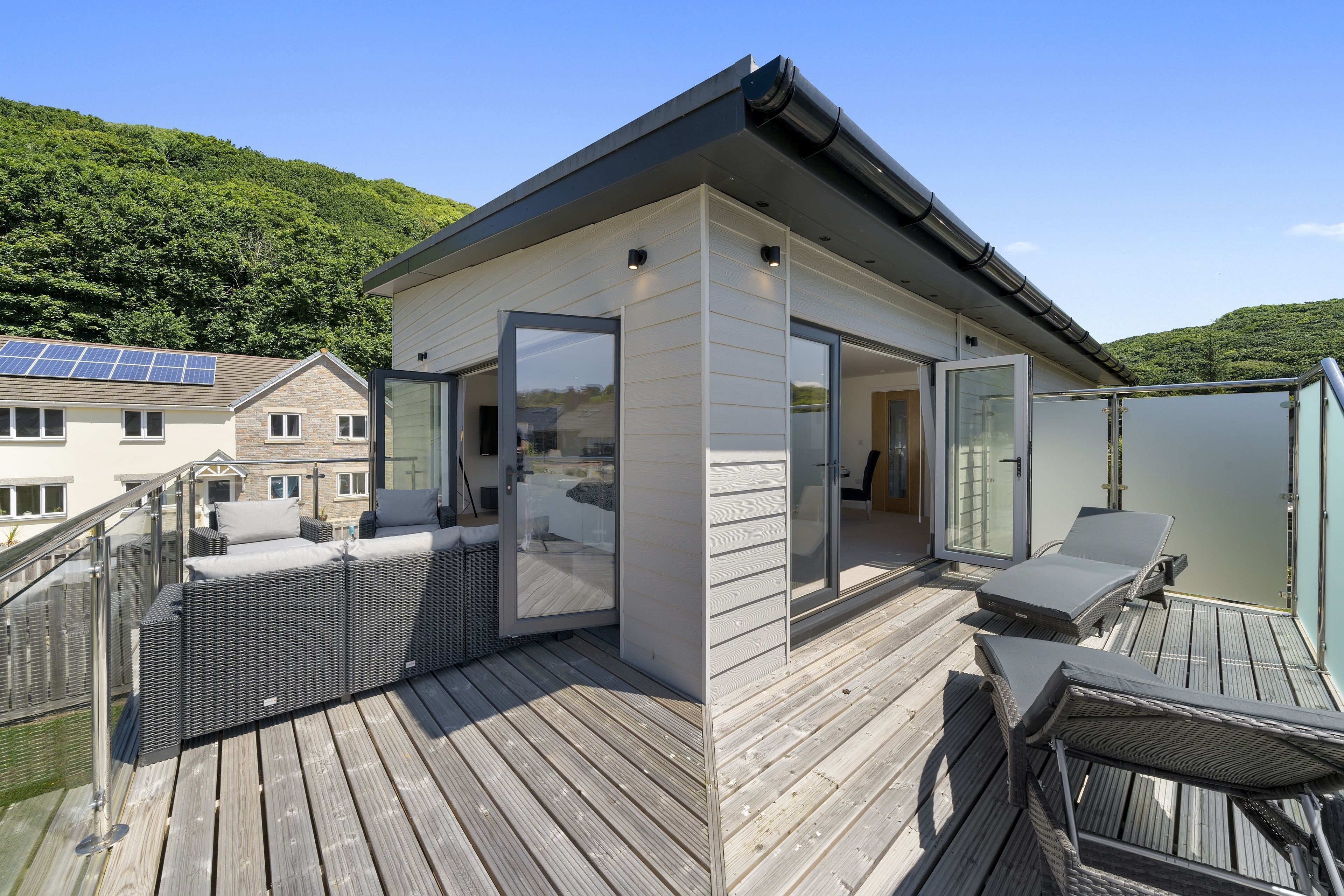 A modern outdoor deck with wicker furniture and glass doors, overlooking a forested hillside.
