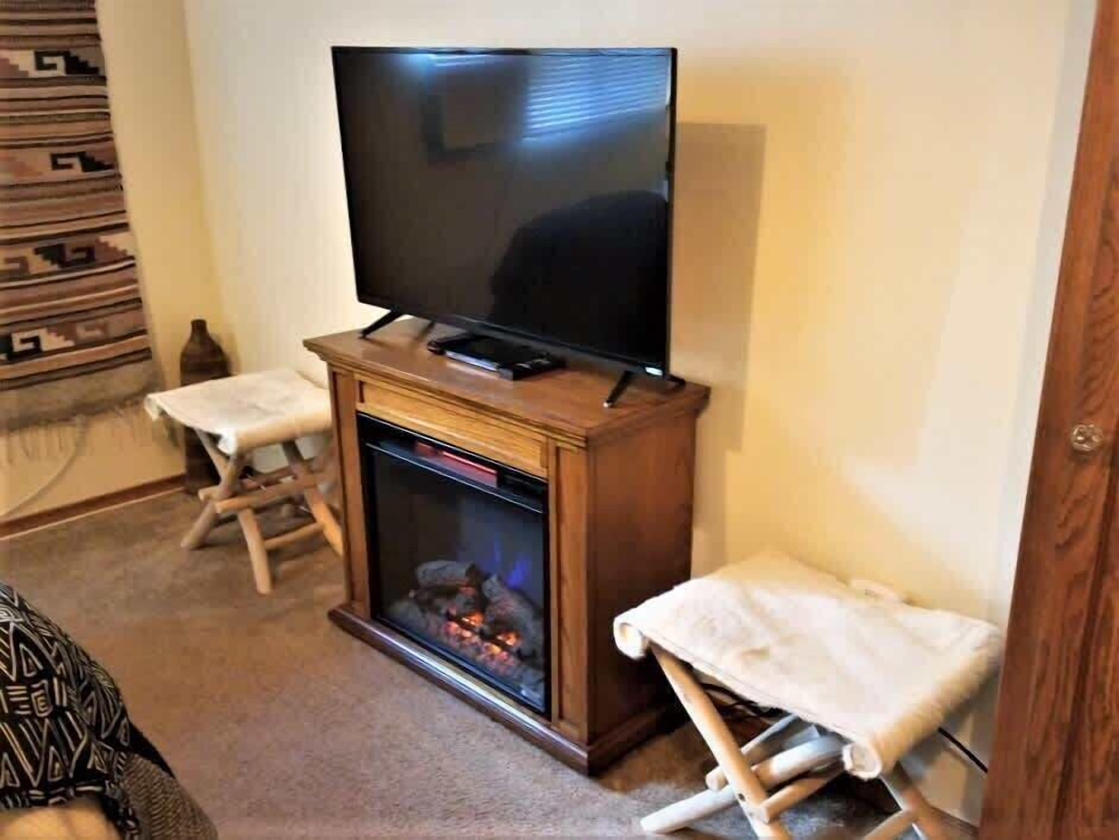 Un soggiorno con una TV a schermo piatto su un supporto in legno, un camino acceso e due tavolini ricoperti da tovaglie bianche.