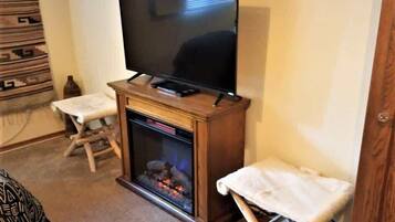 Un soggiorno con una TV a schermo piatto su un supporto in legno, un camino acceso e due tavolini ricoperti da tovaglie bianche.