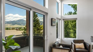Un balcon avec vue sur les montagnes et les arbres, une porte-fenêtre coulissante et un coin salon avec un canapé et un fauteuil en cuir.