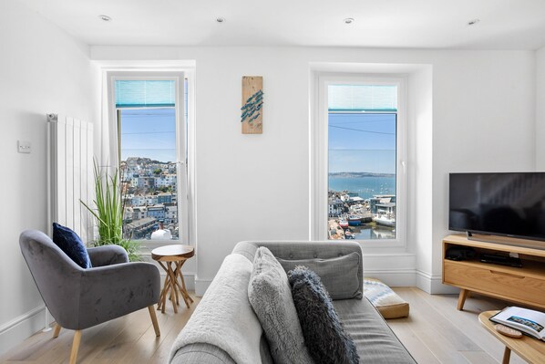 A modern living room with a grey sofa, a grey armchair, a wooden coffee table, and a TV on a wooden stand. There are two windows with a view of a city and the sea.