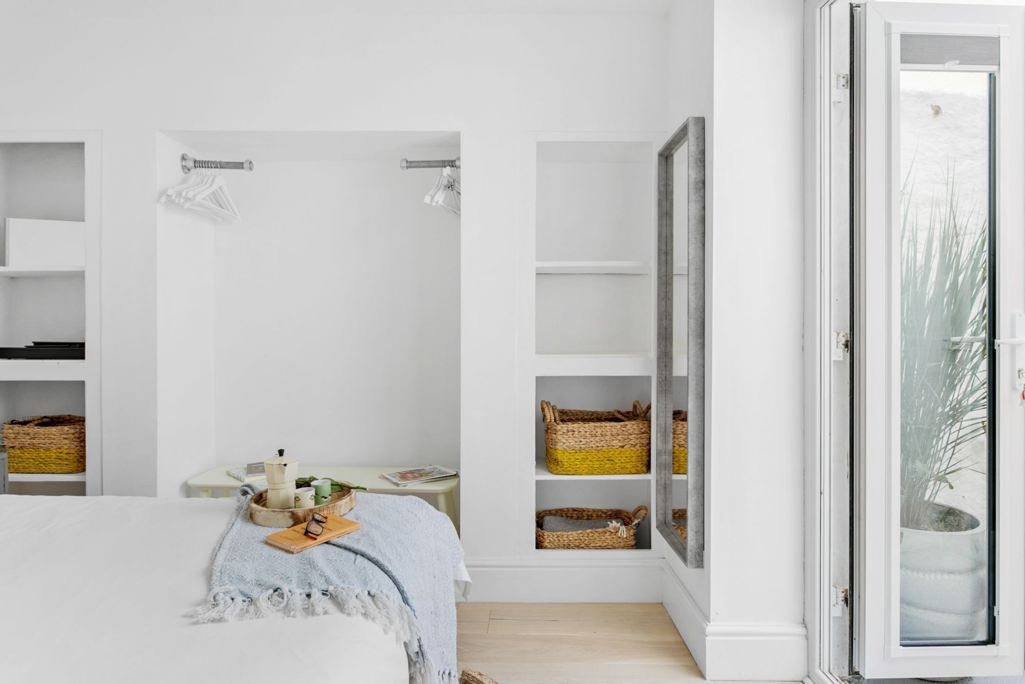 Un dormitorio con cama, una mesita de noche con un libro y gafas, una ventana con vistas a árboles y estanterías empotradas con cestas.