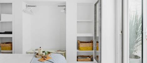 Un dormitorio con cama, una mesita de noche con un libro y gafas, una ventana con vistas a árboles y estanterías empotradas con cestas.