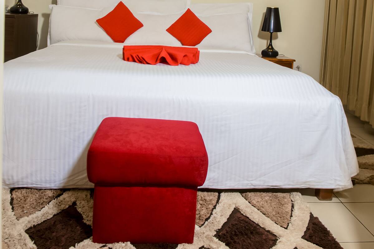A hotel room with a large bed, two bedside tables with lamps, a red ottoman, a patterned rug, and a wall-mounted air conditioner.