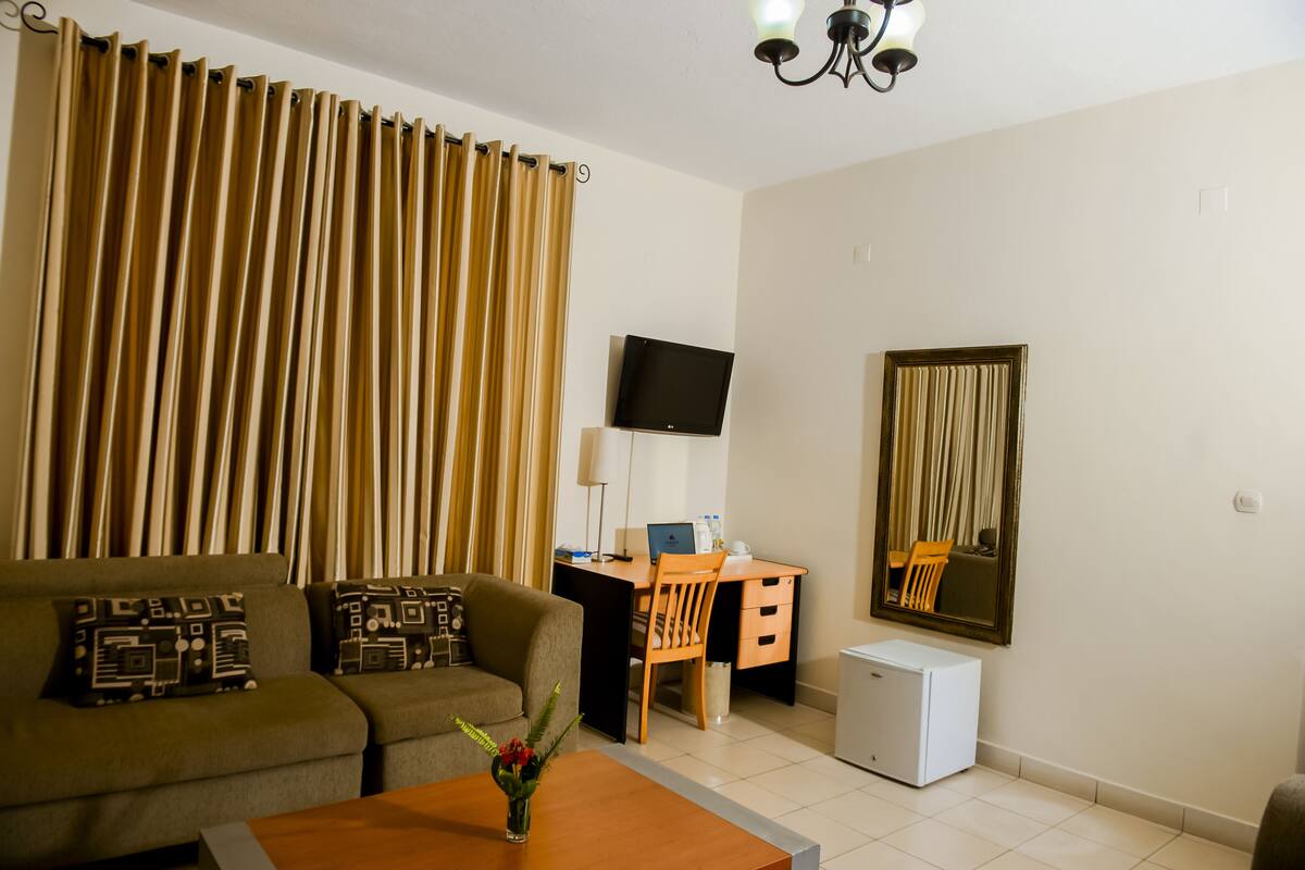 A living room with a sofa, a coffee table, a desk with a computer, a TV, a small fridge, and a mirror on the wall.