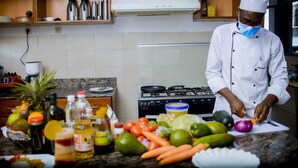 A chef in a white uniform and hat preparing vegetables in a kitchen with a stove, various cooking utensils, and a tiled backsplash. - ONE CLICK HOTEL (Kigali)