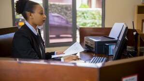 A receptionist at a desk with a laptop, a printer, and a computer monitor. - ONE CLICK HOTEL (Kigali)