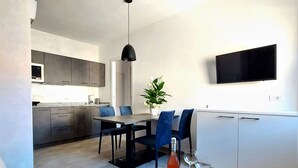 Een moderne woonkamer met een bank, een salontafel met een fles en glazen, een eethoek met een tafel en stoelen, een keuken met kastjes en een magnetron, en een aan de muur bevestigde televisie.