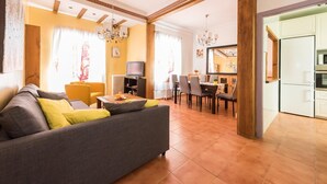 Een woonkamer met een bank, eettafel, stoelen en een televisie. In de achtergrond is een keuken zichtbaar.