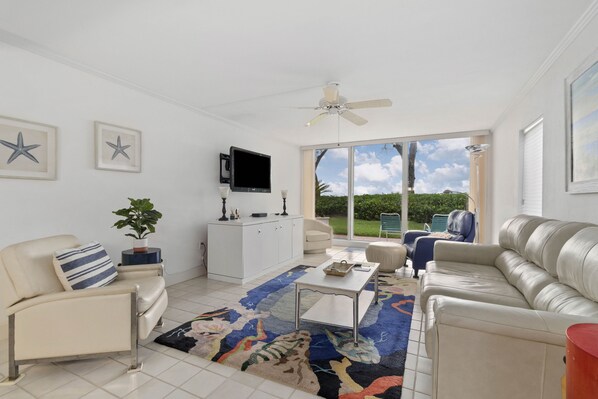 A spacious living room with a white sofa, a coffee table, and a TV mounted on the wall. There is a large window providing a view of the outdoors. - 1st Floor Beachfront Luxury–Ocean Views Included Club Longboat #516 by RVA (Longboat Key)