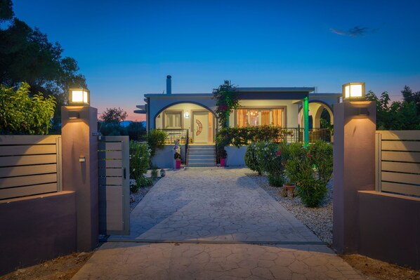 Una casa moderna con un camino pavimentado que conduce a una entrada con portón, rodeada de vegetación.