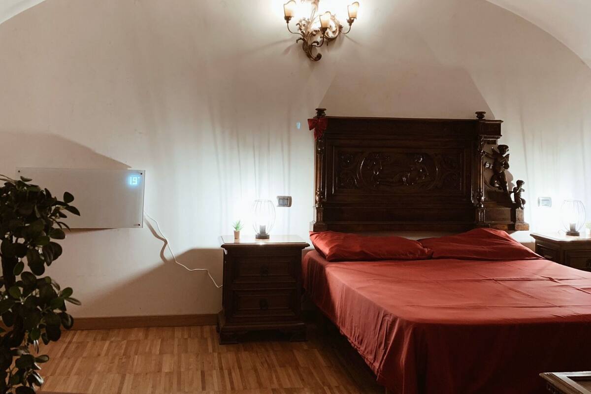Ein Schlafzimmer mit einem hölzernen Bett, einem Nachttisch, einer Stehlampe, einer Zimmerpflanze und einer Wanduhr.