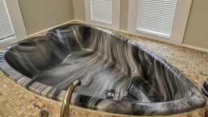 Un grand bain à remous de forme ovale, avec une finition qui rappelle le marbre, installé dans une pièce dont le sol est carrelé et les murs sont blancs.