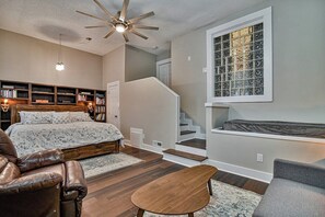 A bedroom with a bed, a leather chair, a wooden table, a ceiling fan, a bookshelf, and a window with a view.