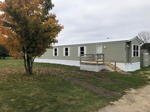 Une maison mobile de plain-pied, dotée d’une véranda, d’un petit abri de jardin et d’un arbre aux feuilles d’automne.