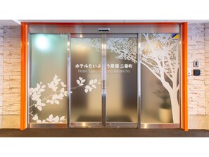 Entrada de hotel con diseño moderno, destacando una puerta de vidrio con motivos de árbol y ramas, y señalización bilingüe en japonés e inglés.