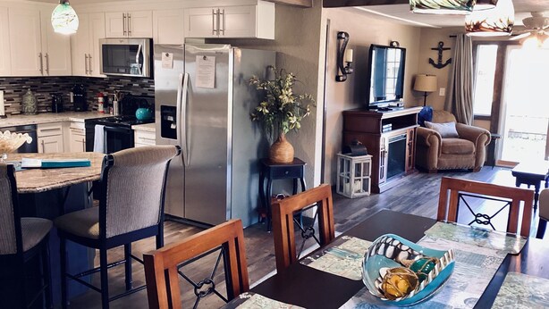 Una cocina moderna con electrodomésticos de acero inoxidable, un comedor con sillas y mesa de madera, y una sala de estar acogedora con televisor y asientos cómodos.