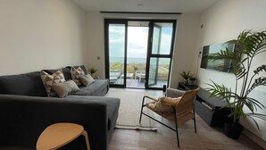 A modern living room with a grey sofa, a wicker chair, and a wooden table. There is a large window with a view of the outdoors. - Beautiful Apartment on Ramsgate Sea-front (Ramsgate)