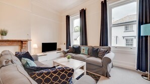 Ein Wohnzimmer mit einer Sofagarnitur, einem Couchtisch, einem Flachbildfernseher, einem Kamin und großen Fenstern mit Vorhängen.