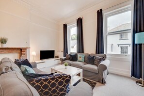 Ein Wohnzimmer mit einer Sofagarnitur, einem Couchtisch, einem Flachbildfernseher, einem Kamin und großen Fenstern mit Vorhängen.
