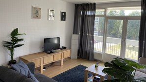 Ein modernes Wohnzimmer mit einem Flachbildfernseher auf einem Holz-TV-Möbel, einem grauen Sofa, einem Couchtisch und einem großen Fenster mit Vorhängen.