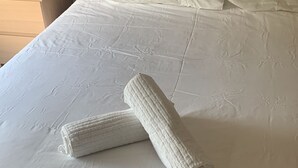 Ein ordentlich bezogenes Bett mit weißen Leinen, ein gerolltes Handtuch und eine Nachttischlampe auf einem Nachttisch aus Holz mit einer kleinen Pflanze.