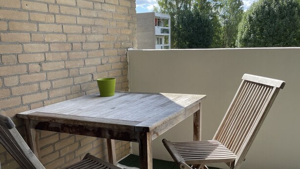 Eine kleine Terrasse im Freien mit einem Holztisch und zwei Stühlen, einer grünen Schüssel und einem gemusterten Teppich.
