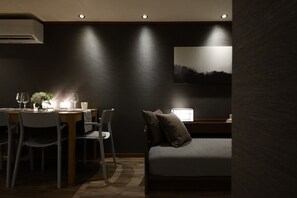 Una habitación tenuemente iluminada con una mesa de comedor dispuesta para dos, una cama individual con un almohón y un cuadro en la pared.