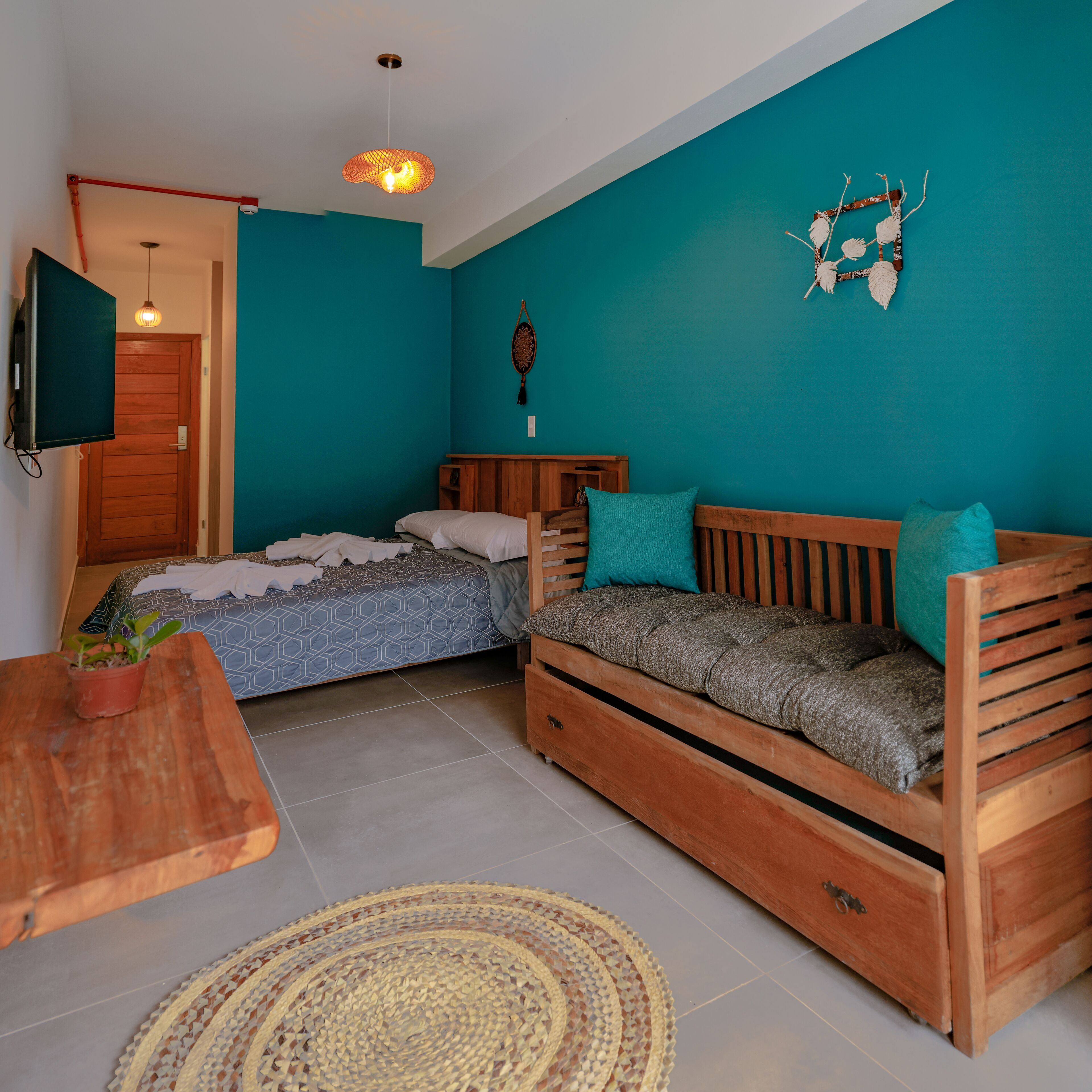 A compact room with teal walls, a wooden sofa, a bed with patterned bedding, a small wooden table with a plant, and a television mounted on the wall.