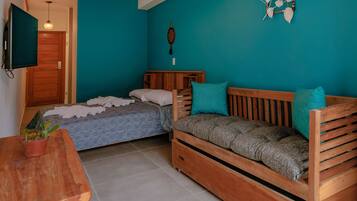 A compact room with teal walls, a wooden sofa, a bed with patterned bedding, a small wooden table with a plant, and a television mounted on the wall.