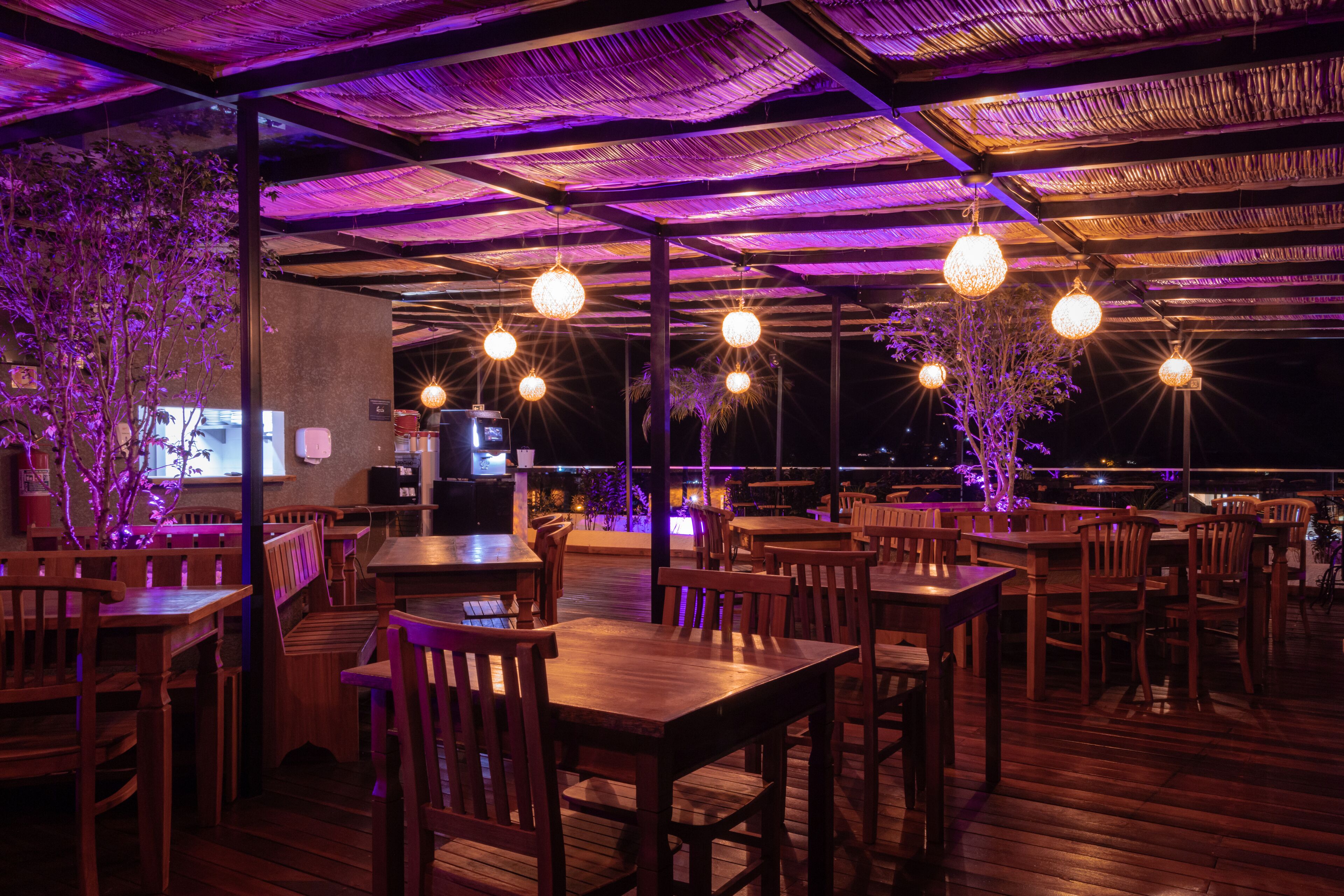 Restaurante ao ar livre com mesas e cadeiras de madeira, luminárias esféricas penduradas e vista para a cidade à noite.