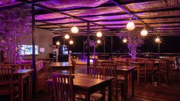 Restaurante ao ar livre com mesas e cadeiras de madeira, luminárias esféricas penduradas e vista para a cidade à noite.