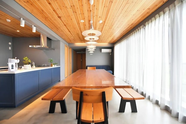 A modern kitchen with a wooden ceiling, a long dining table with benches, and a view of the outdoors through large windows.