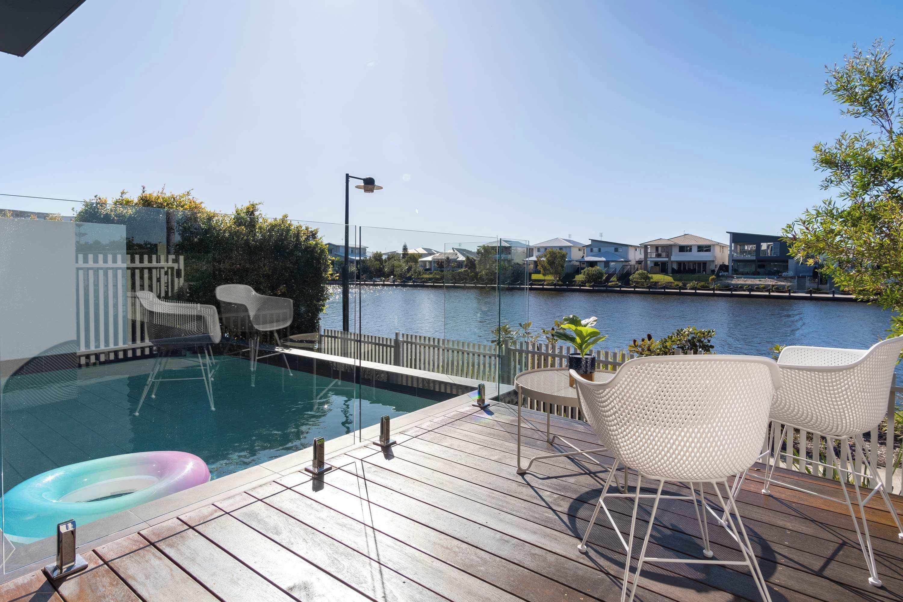 A pool area with a wooden deck, white wicker chairs, and a view of houses across the water.