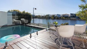 A pool area with a wooden deck, white wicker chairs, and a view of houses across the water. - Luxury Lakeside Living at Birtinya (Birtinya)