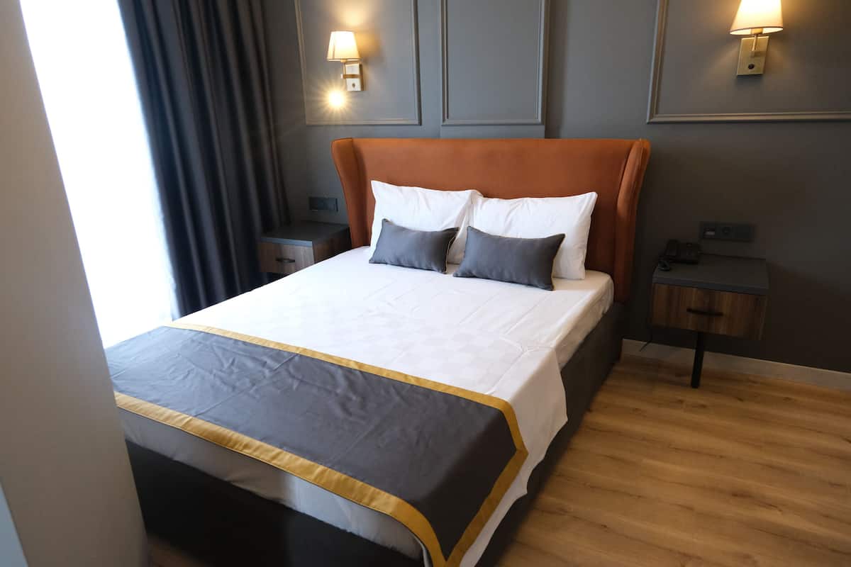 Una cama impecablemente arreglada con colcha gris y dorada, dos almohadas y un cabecero de madera. Hay dos mesitas de noche con lámparas y un buró con una lámpara. La habitación tiene suelo de madera y una pared gris con un cuadro enmarcado.