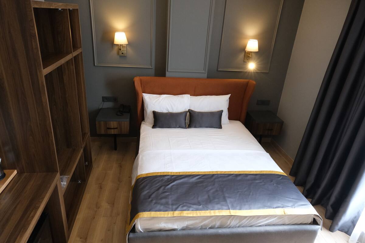 Una cama bien hecha con edredón gris y blanco, cabecera de madera y mesitas de noche con lámparas.