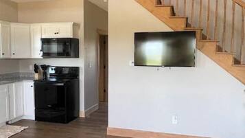 A spacious living area with a wooden staircase, a kitchen with white cabinets, and a flat-screen TV mounted on the wall.