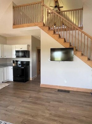 A spacious living area with a wooden staircase, a kitchen with white cabinets, and a flat-screen TV mounted on the wall.