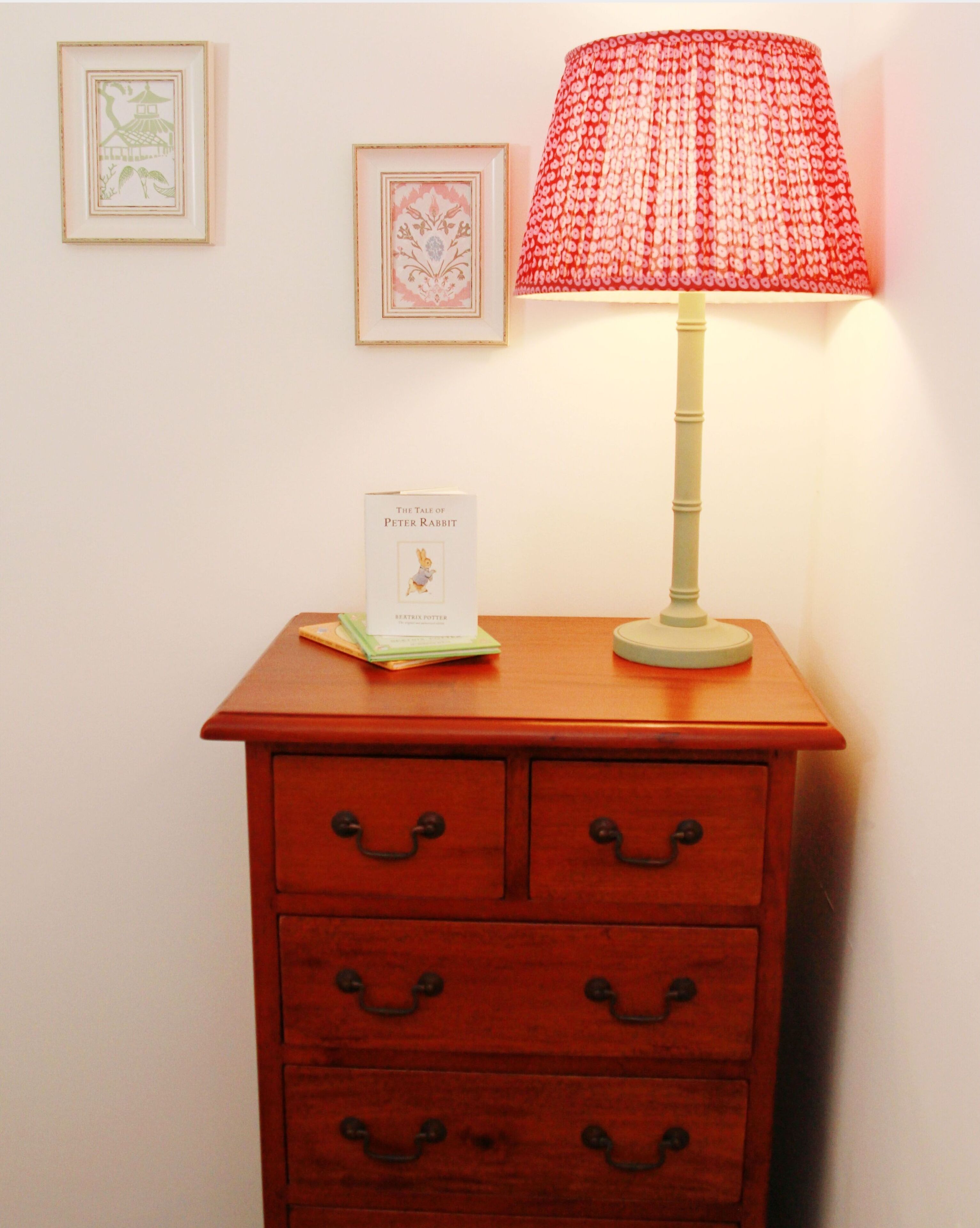 A wooden nightstand with a lamp, a book titled "The Tale of Peter Rabbit," and framed pictures on the wall.