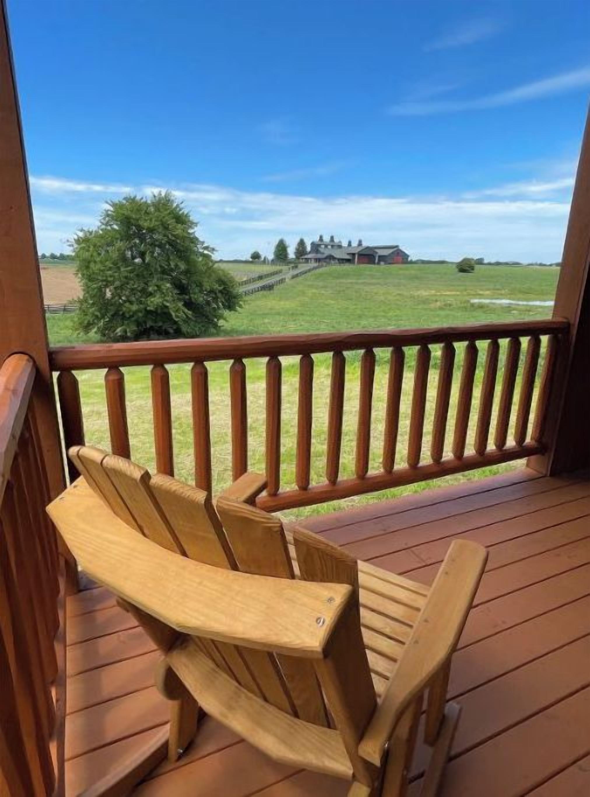 Une terrasse en bois avec une chaise en bois et une rambarde, donnant sur un champ verdoyant et un bâtiment situé au loin.